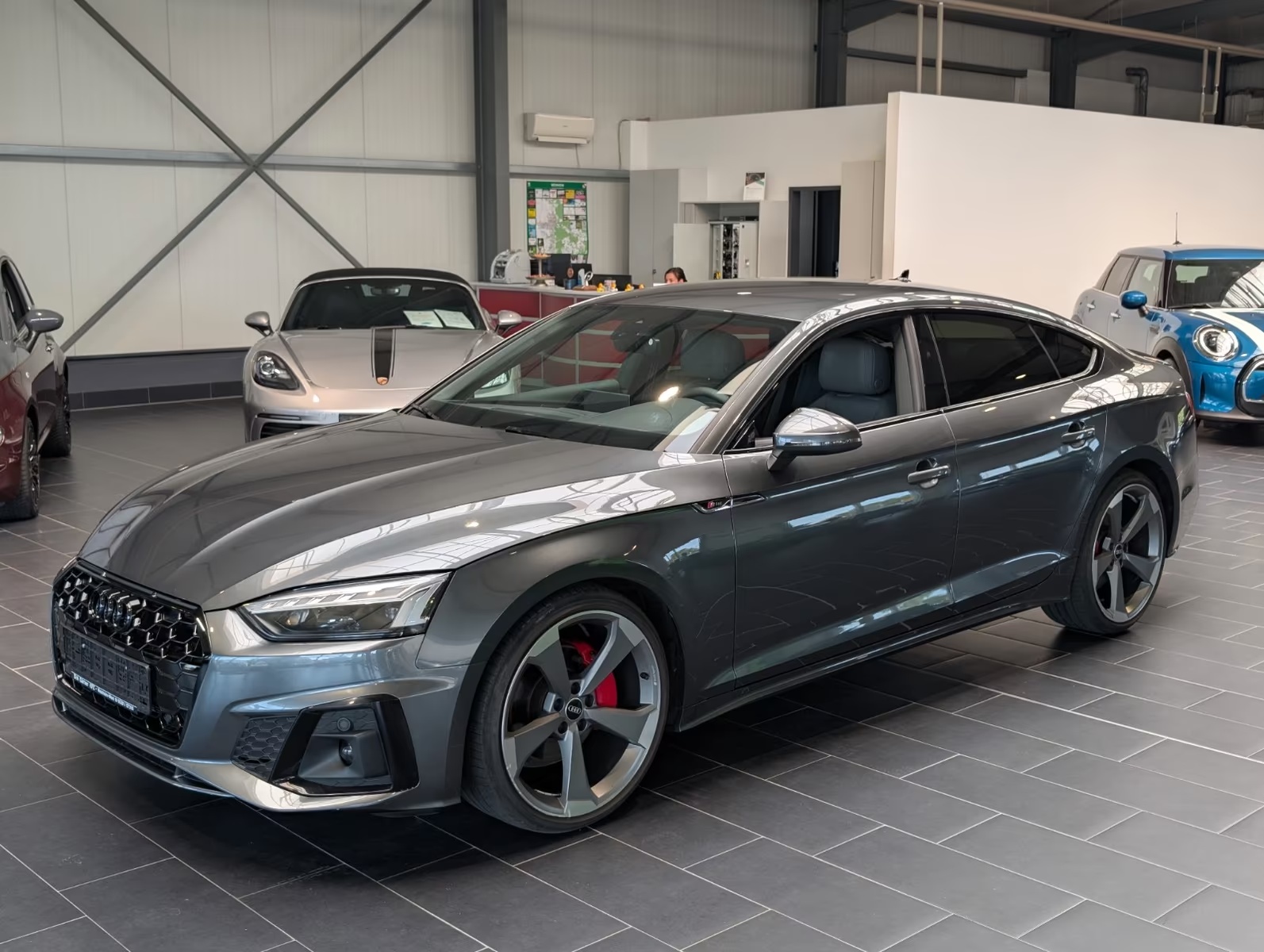 Audi A5 Sportback S line in Daytona Grey metallic, front three-quarter view from the driver side inside a dealership showroom.