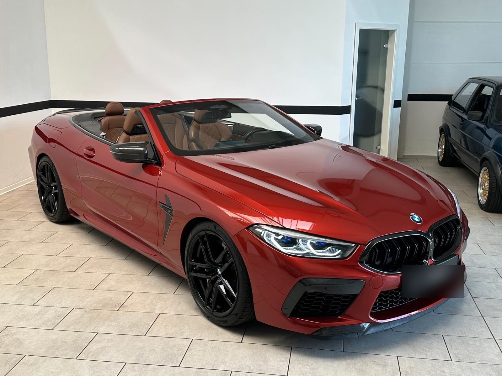 Front three-quarter view of the BMW M8 Cabrio from the passenger side, showing the carbon mirror caps and cognac interior.