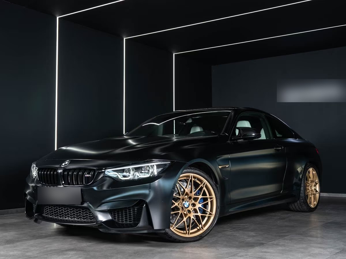 BMW M4 Competition Coupé in Frozen Black, front three-quarter view with gold M double-spoke wheels in a dark showroom.