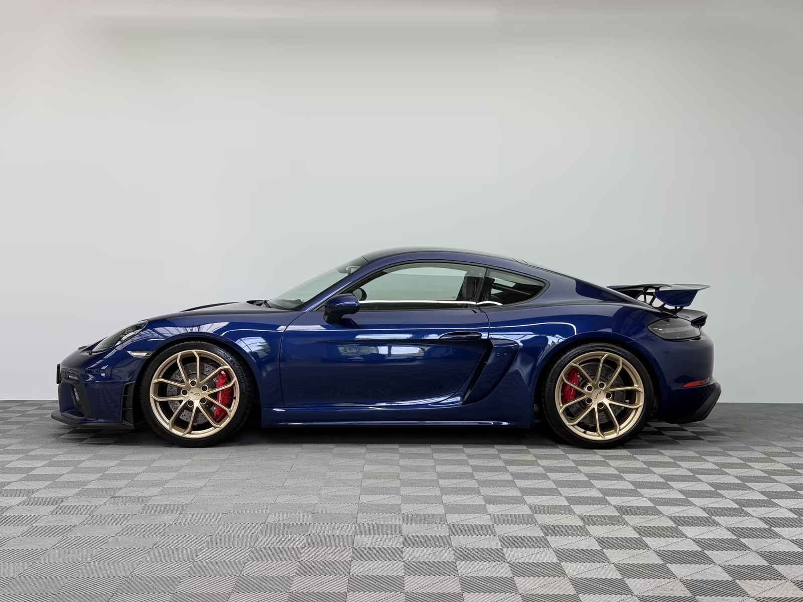 Side profile of the blue Porsche 718 Cayman GT4 showing the fixed rear wing, side air intake and Aurum alloy wheels with red brake calipers.