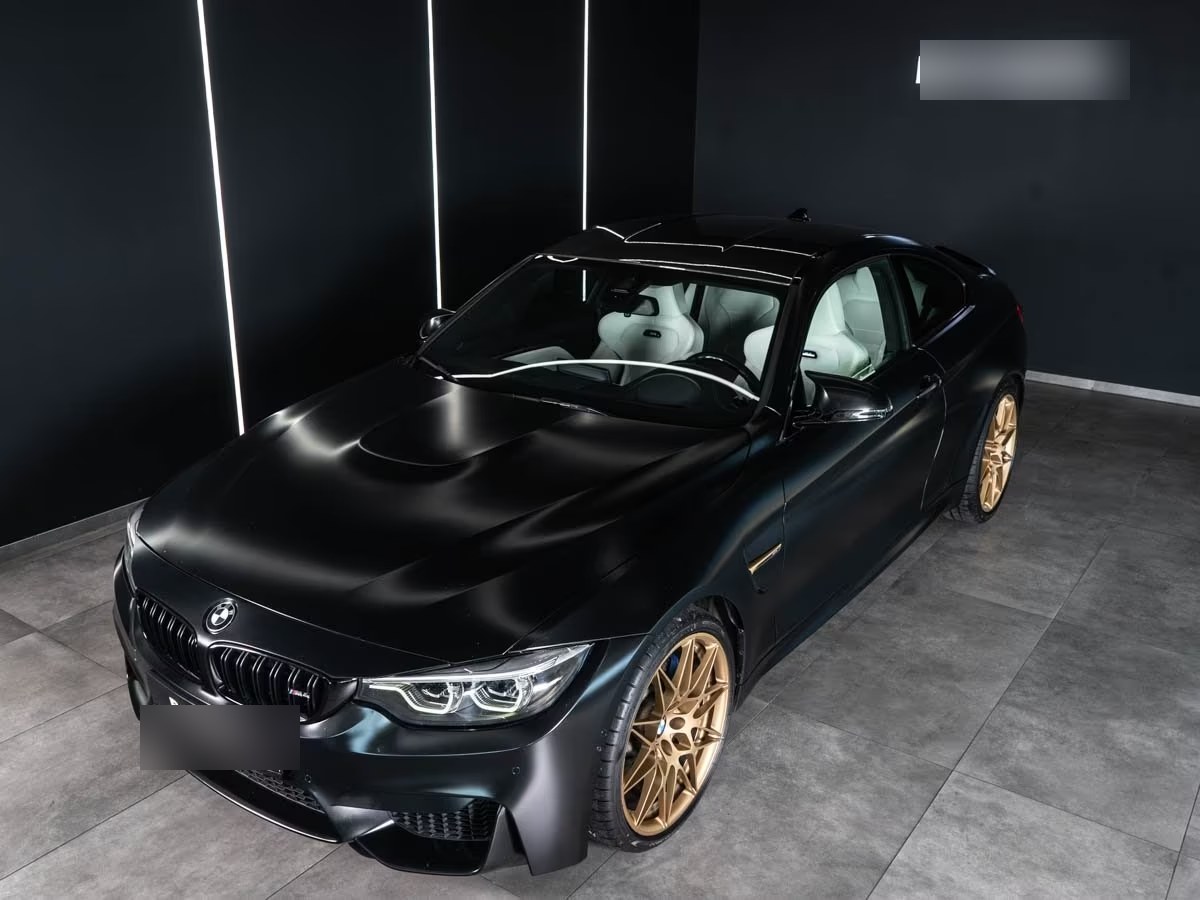 Elevated front three-quarter view of the Frozen Black BMW M4 showing the bonnet power dome and white leather interior.
