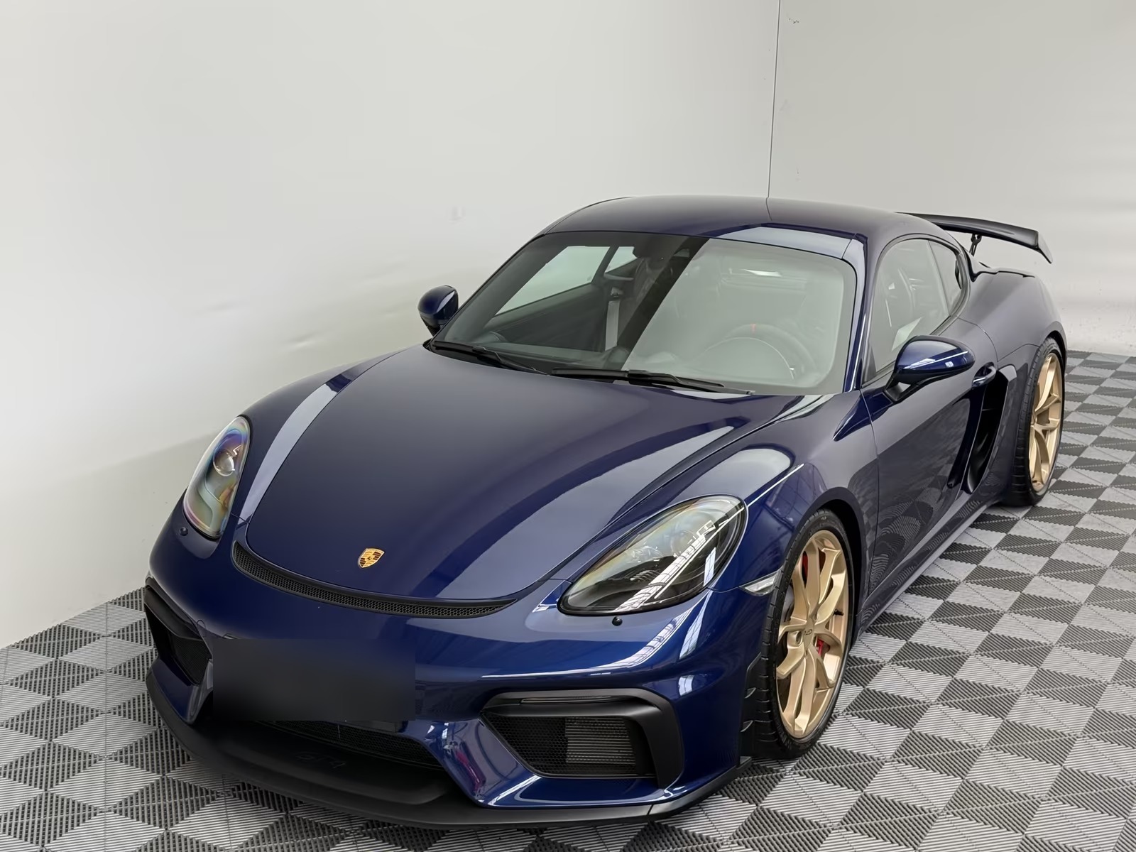Porsche 718 Cayman GT4 in Gentian Blue metallic, front three-quarter view from the driver side showing the GT4 front bumper and gold alloy wheels.