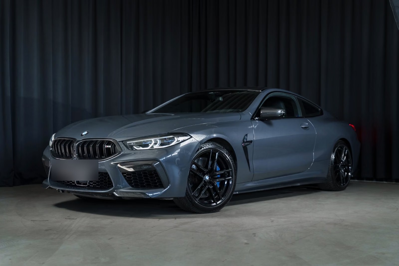 BMW M8 Coupé in Brands Hatch grey metallic, front three-quarter view against a dark studio backdrop.